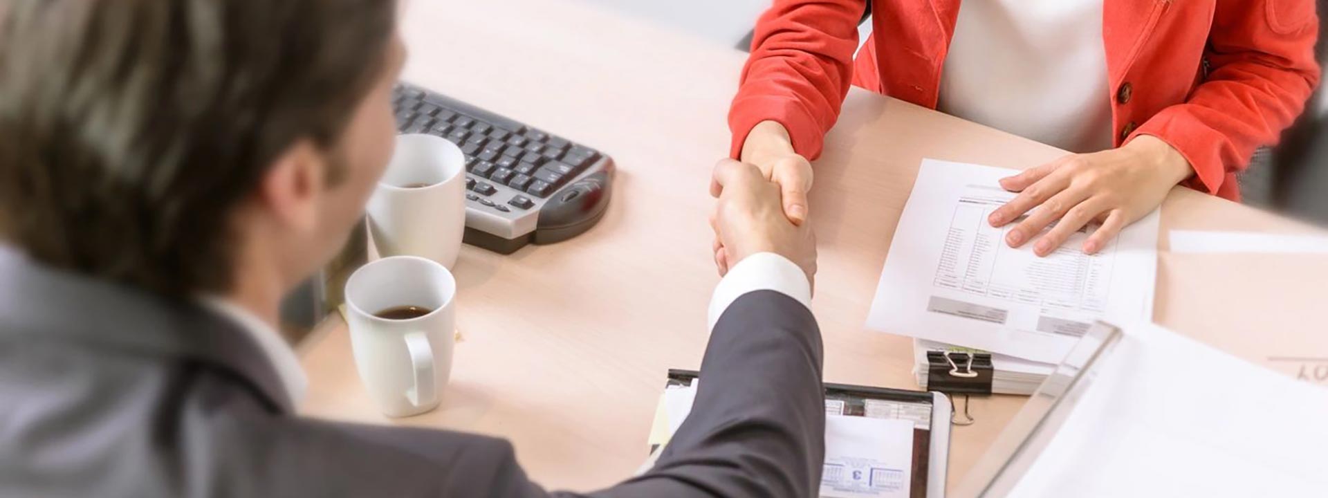 Two people shaking hands over a desk with cups of coffee.